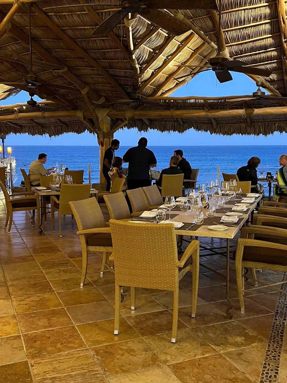 Outdoor ocean-view restaurant with traditional thatched roof and elegant dining setup.