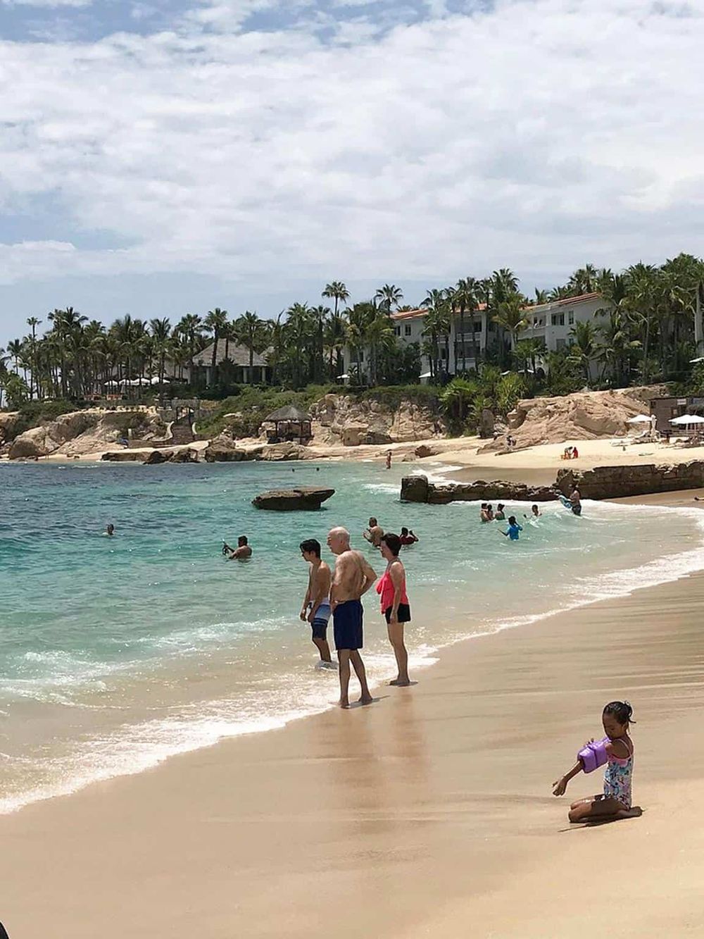 Relaxing beach scene with palm trees, hotel resorts, and families enjoying water activities and sandy shores.