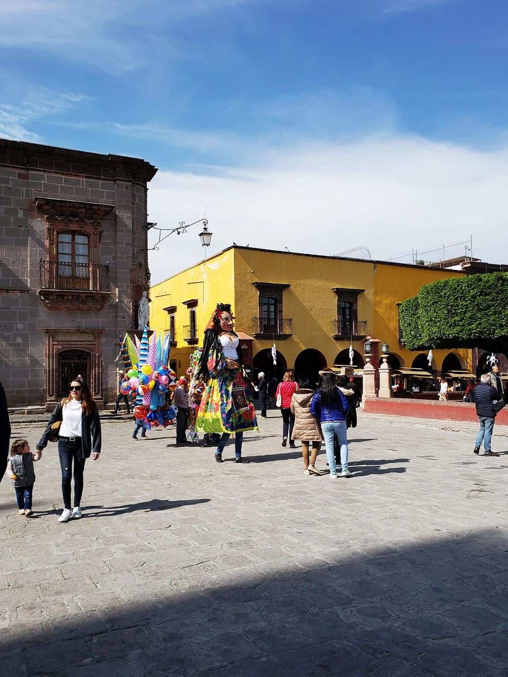 Colorful street scene featuring a giant traditional doll, vibrant buildings, and tourists in a historic plaza.
