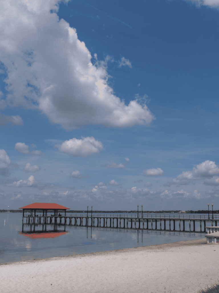 Tranquil lakeside pier with gazebo and beach under blue sky with clouds, ideal for scenic summer outings.