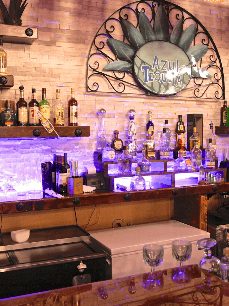 Brightly lit bar with tequila bottles, decorative wall sign, and illuminated shelving at Azul Tequila restaurant.