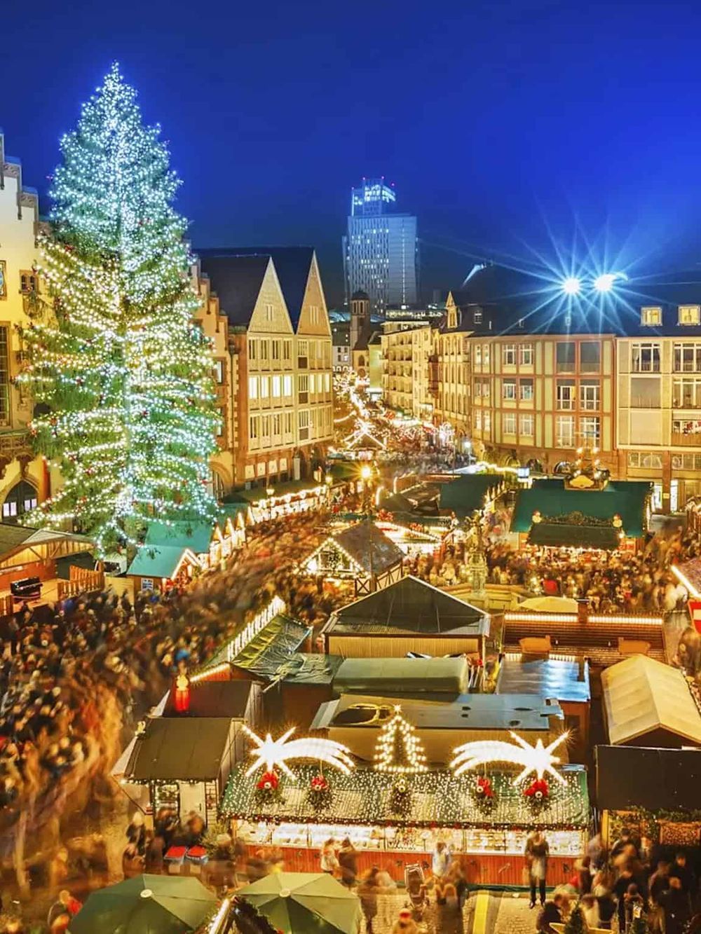 Festive Christmas market with illuminated tree, bustling crowd, and city skyline in the background.