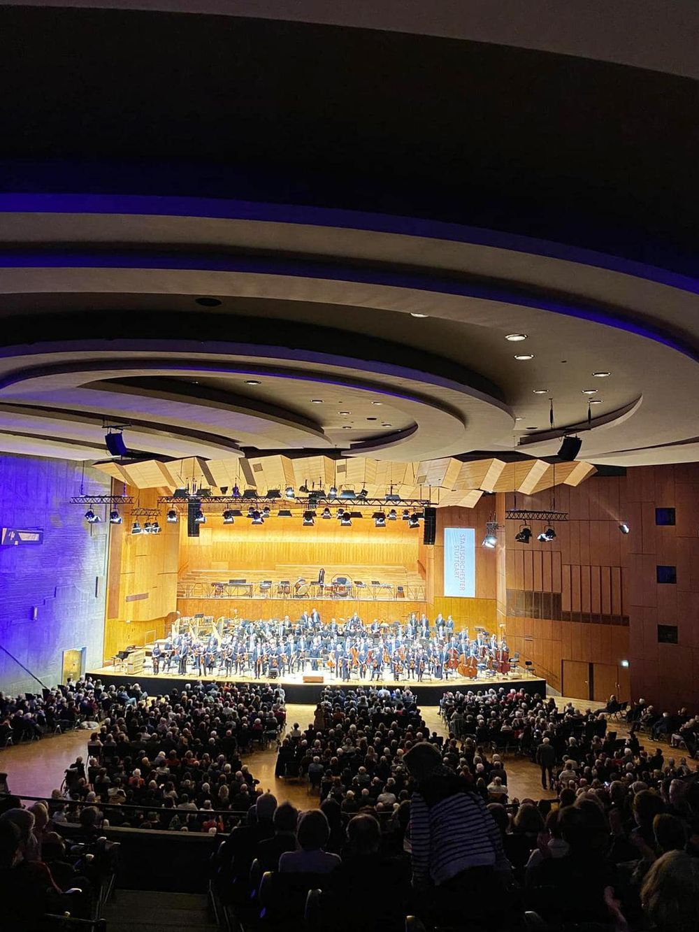 A concert hall filled with an orchestra performing on stage, with an audience seated in front.