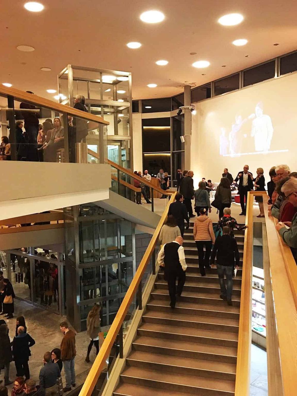 Modern conference center interior with people attending an event, featuring stairs and a large projection screen.