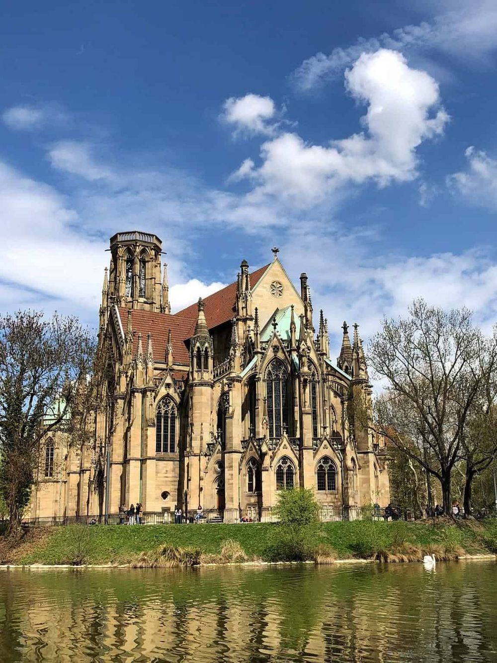 Imposing Gothic-style church with intricate architecture near water, under a bright blue sky with scattered clouds.