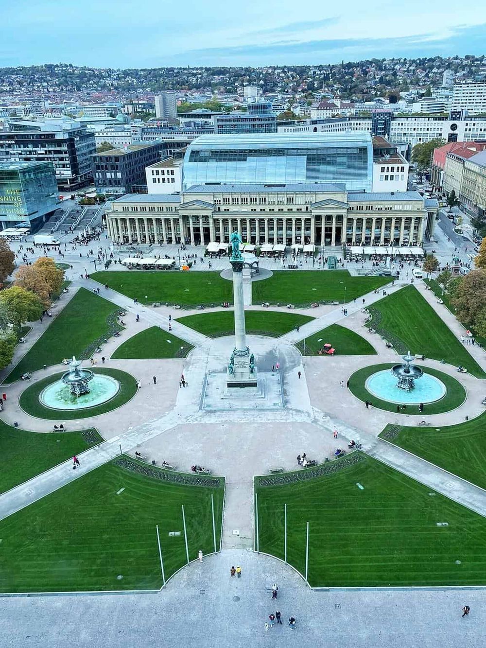 Aerial view of a historic city square with green lawns, fountains, and a prominent statue, showcasing vibrant city life and architecture.
