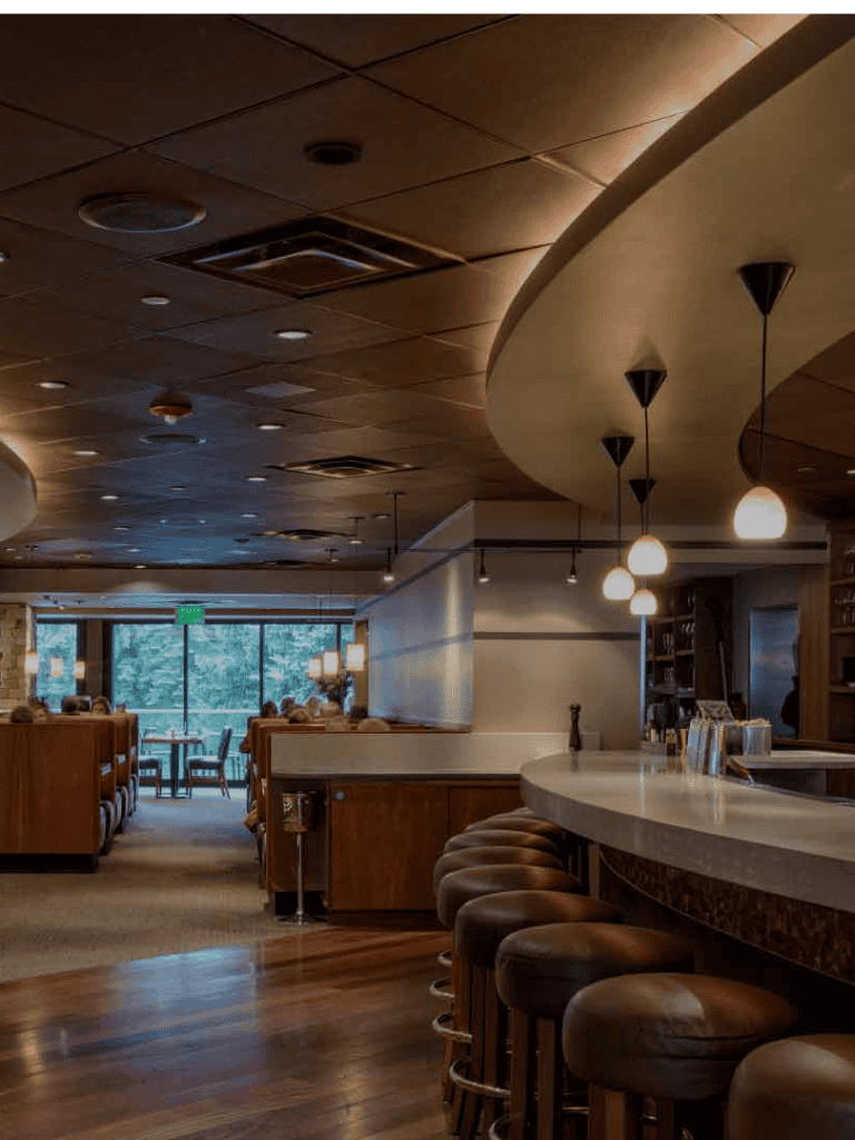 Cozy restaurant interior with bar seating, warm lighting, and large windows overlooking greenery.