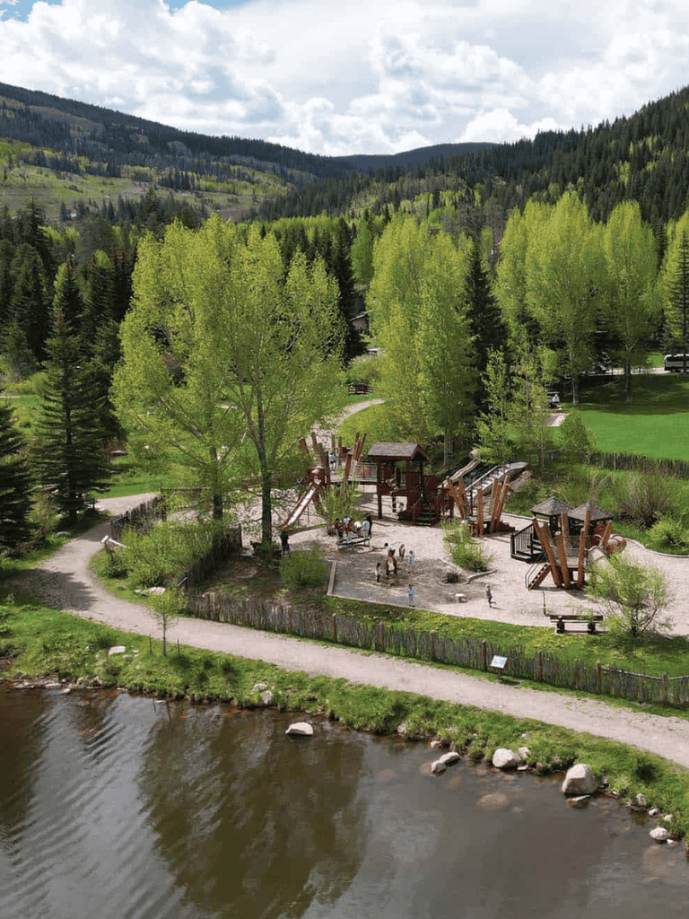 Lush green forested mountainside with outdoor playground near river, scenic nature setting.