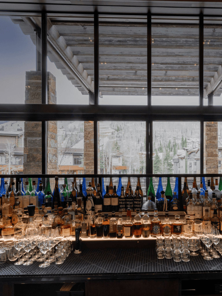 Elegant bar with diverse spirits and glassware, snowy mountain view, cozy indoor setting.