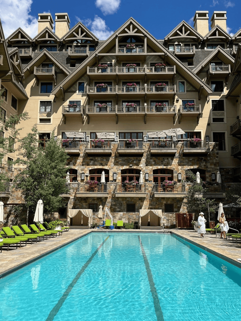 Luxurious hotel building with outdoor pool, sun loungers, and garden area in mountain resort setting.