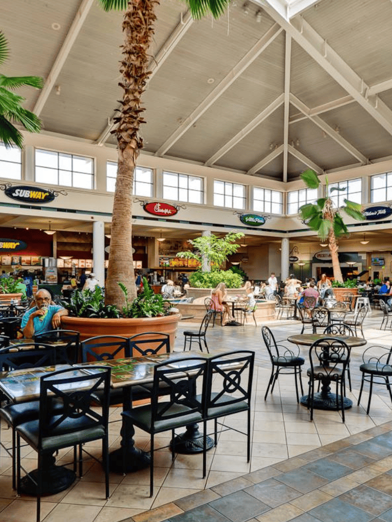 Bright indoor mall food court with palm trees and various restaurant signs, including Subway and Chick-fil-A.