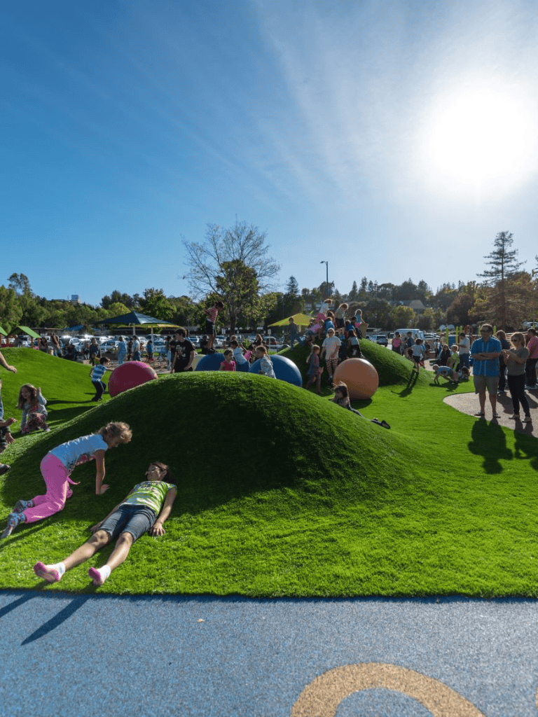 Colorful outdoor park with children playing on grassy hills and people enjoying sunny day, family-friendly recreational space.