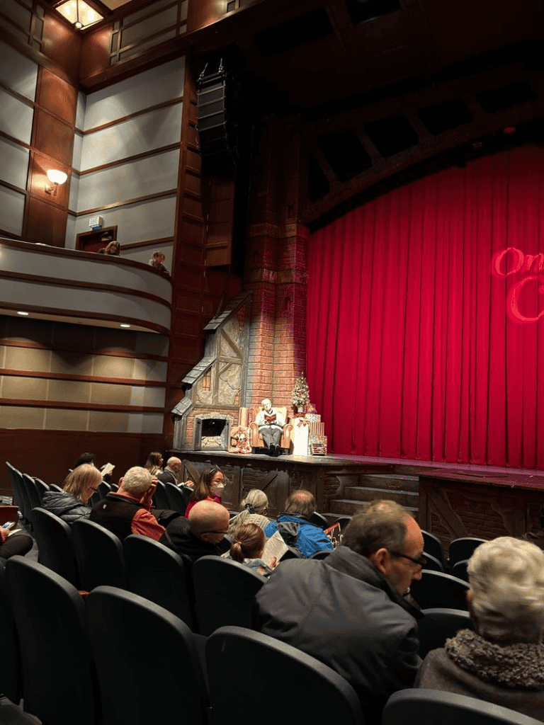 A theater audience watches a holiday-themed performance on stage with red curtains.