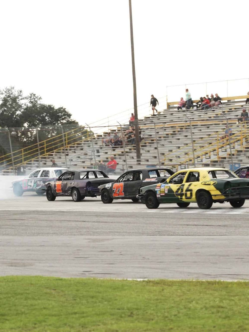 Stock car racing event at an outdoor track with vintage cars and spectators on bleachers.