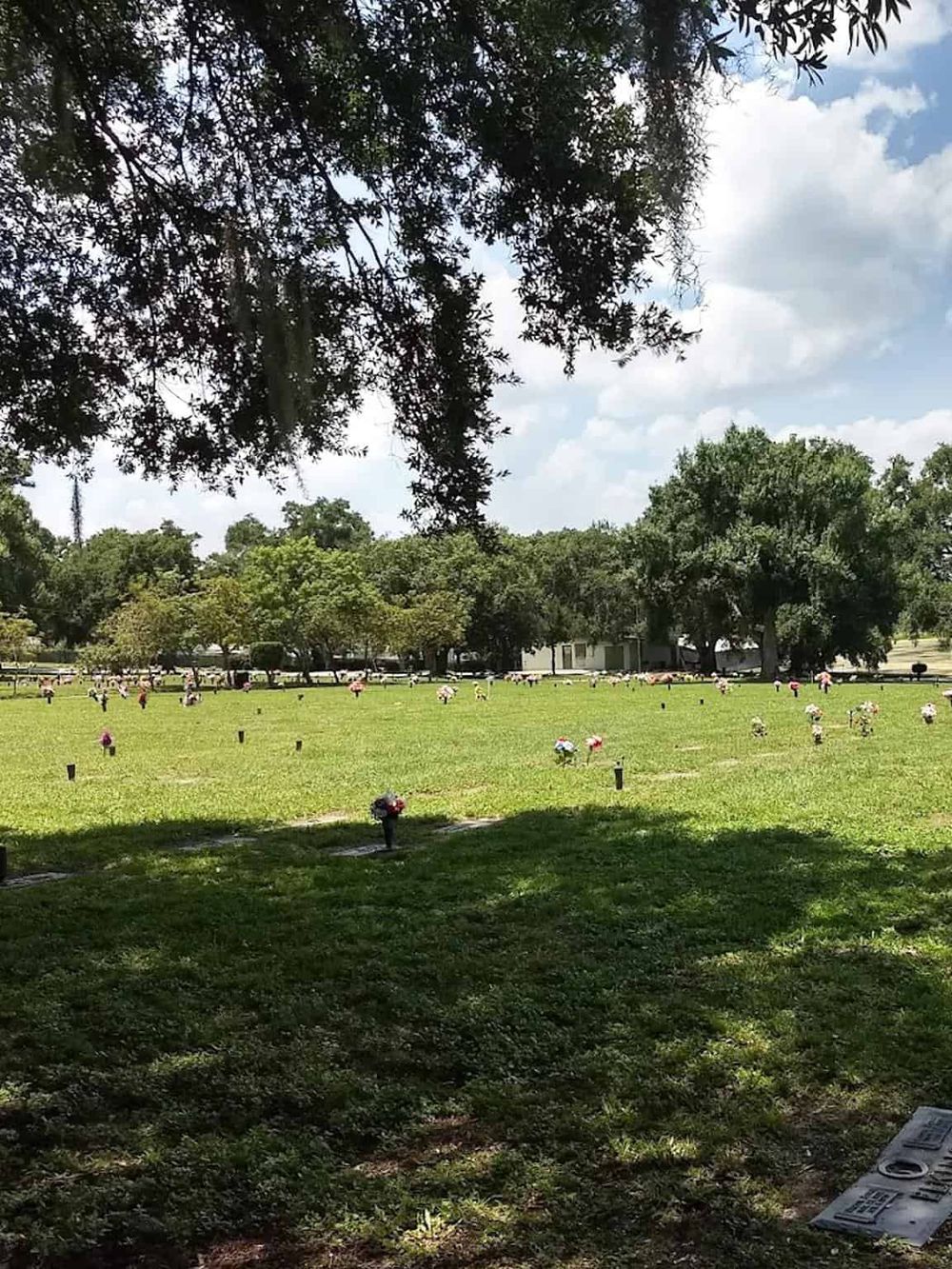 Flowers memorial in a lush green cemetery with trees, shaded areas, clear sky, and a peaceful atmosphere.