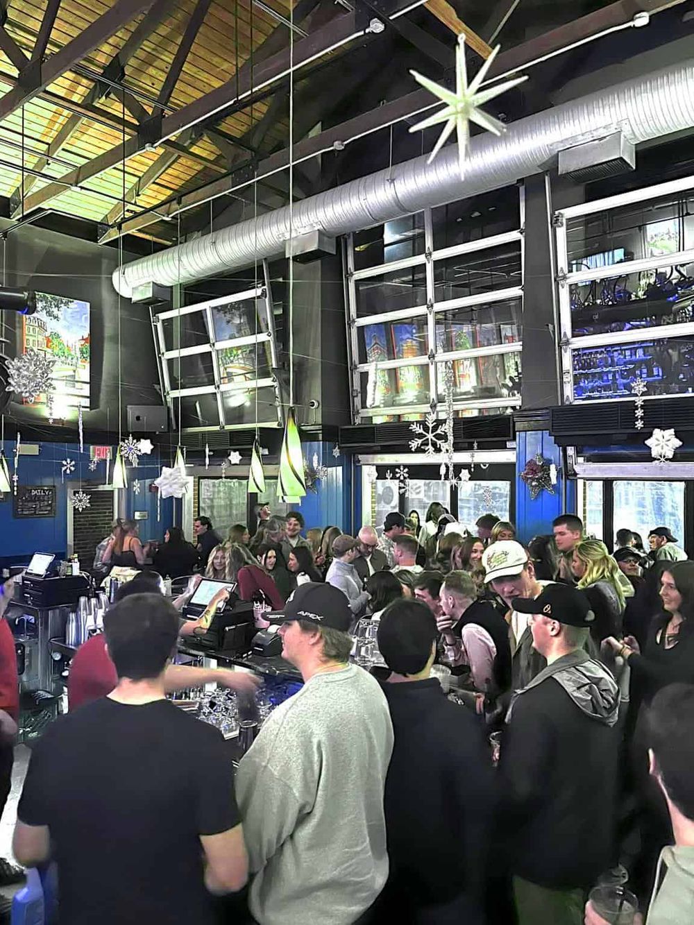 Crowded bar with people enjoying drinks, modern industrial decor, and festive holiday decorations.