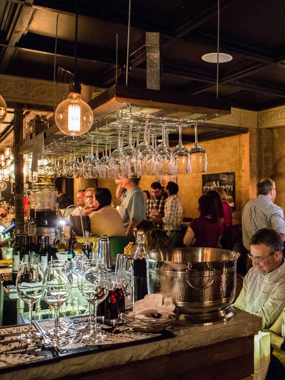 Upscale bar with hanging wine glasses and customers enjoying drinks at a lively restaurant.