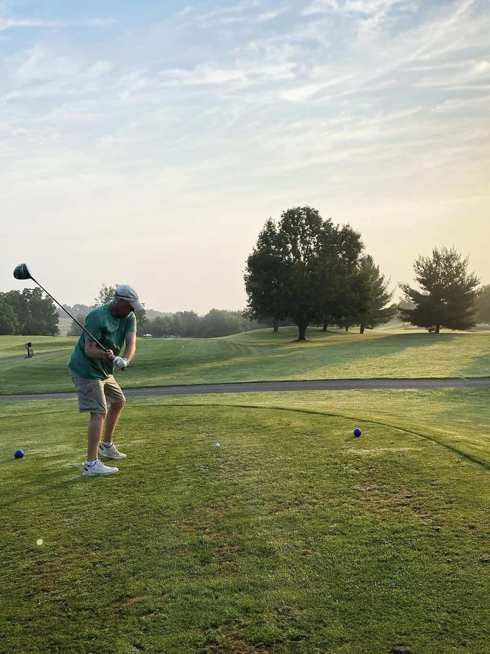 Golf player teeing off at a scenic golf course during sunset with lush green landscape.