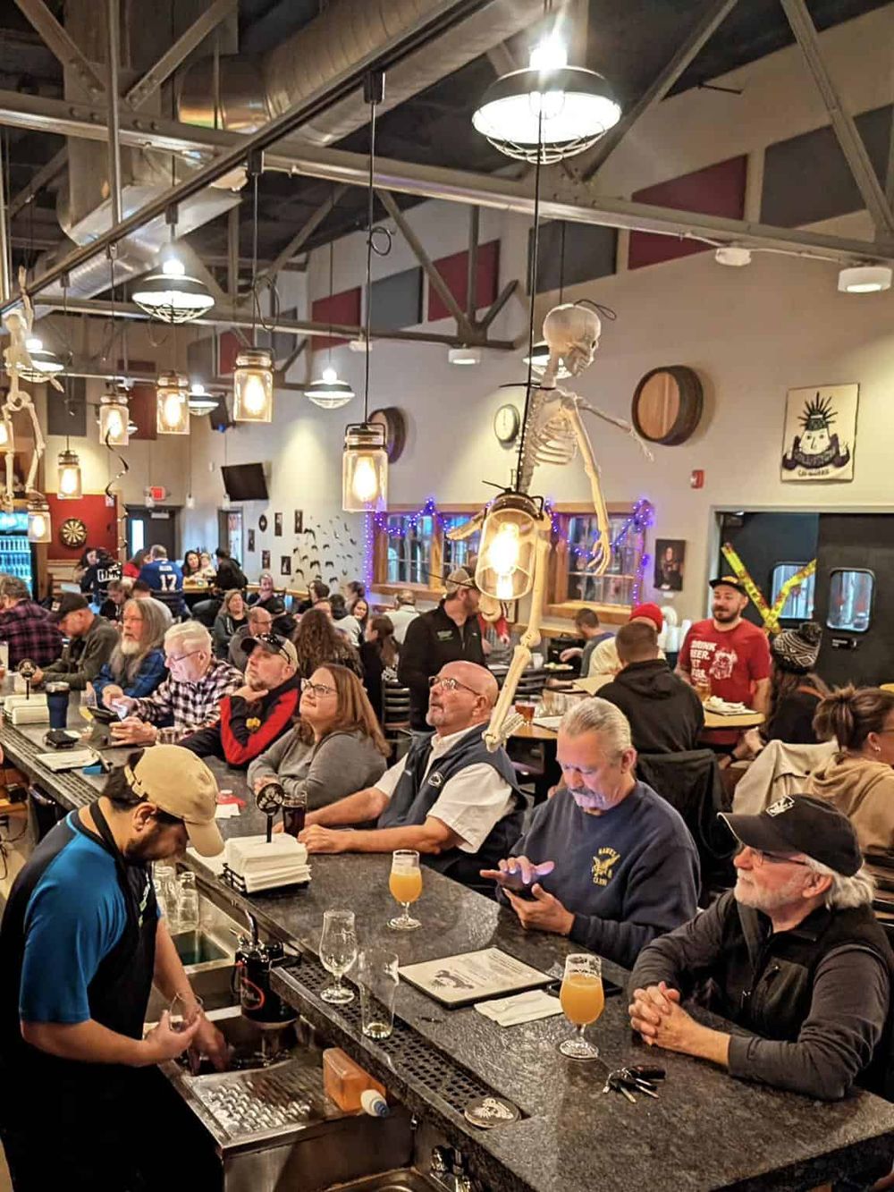 Skeleton Halloween decoration hanging above bar with people enjoying drinks inside a lively restaurant or bar.