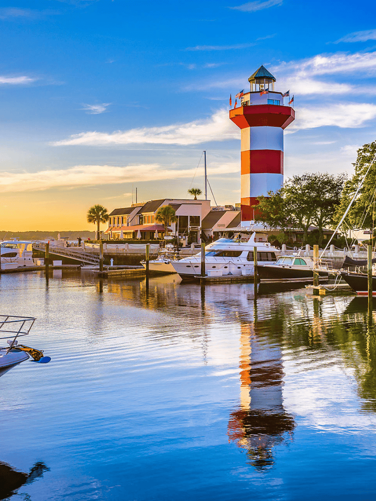 Colorful lighthouse at Marina with boats and waterfront buildings during sunset, scenic harbor view.