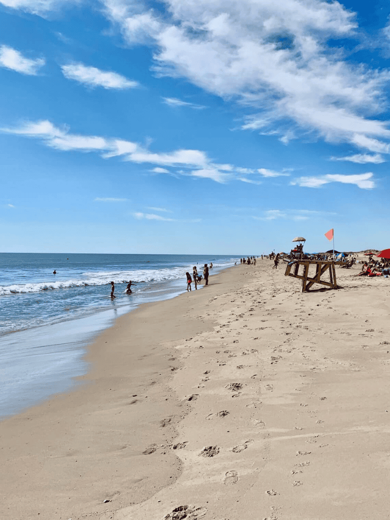 Bright blue sky over sandy beach with footprints, beachgoers, and ocean waves, ideal for beach vacation planning.