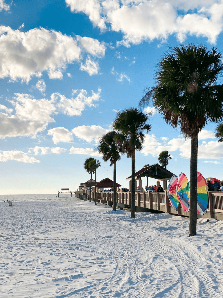 Bright beach scene with palm trees, colorful umbrellas, and a pier under a partly cloudy sky, perfect coastal getaway.