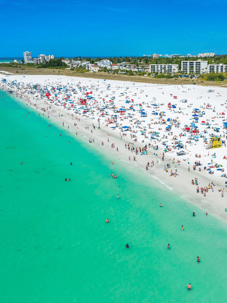 Bright sunny beach scene with turquoise waters, crowded sandy shoreline, and lively atmosphere.