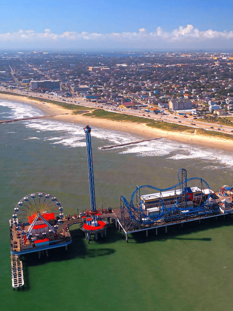 Aerial view of a coastal amusement park with a roller coaster and Ferris wheel by the beach.