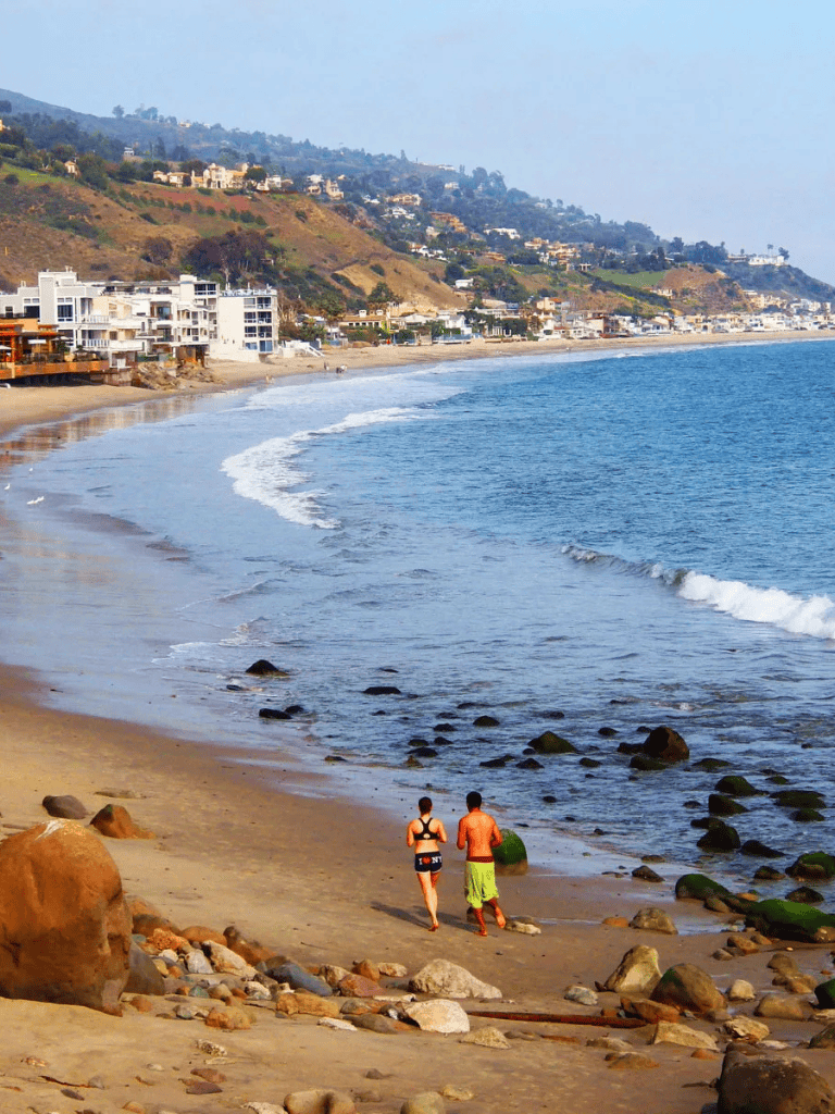 Relaxing beach scene with couple walking on sandy shore, ocean waves, hillside homes in distance, and clear sky.