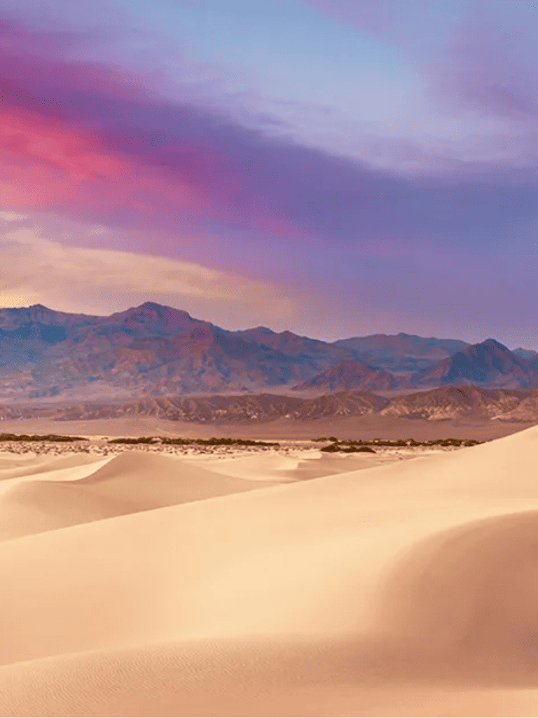 Endless desert sand dunes with mountain backdrop and colorful sky, stunning desert landscape, travel, adventure, outdoor exploration.