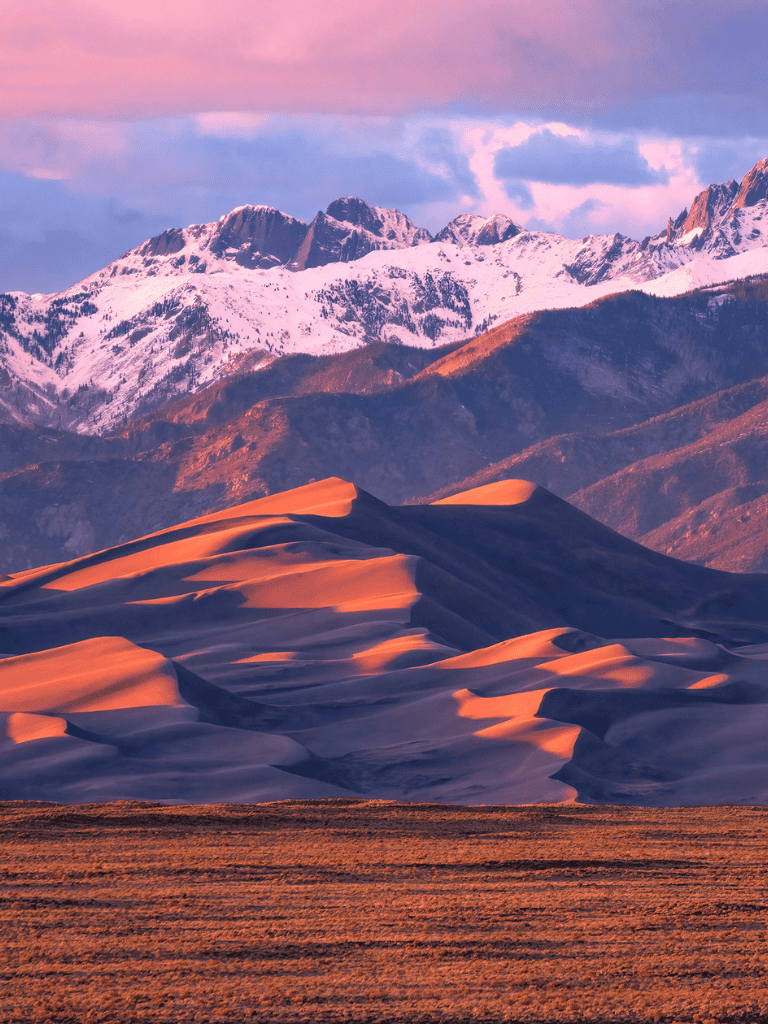 Vast sand dunes with snow-capped mountains in the background during sunset.
