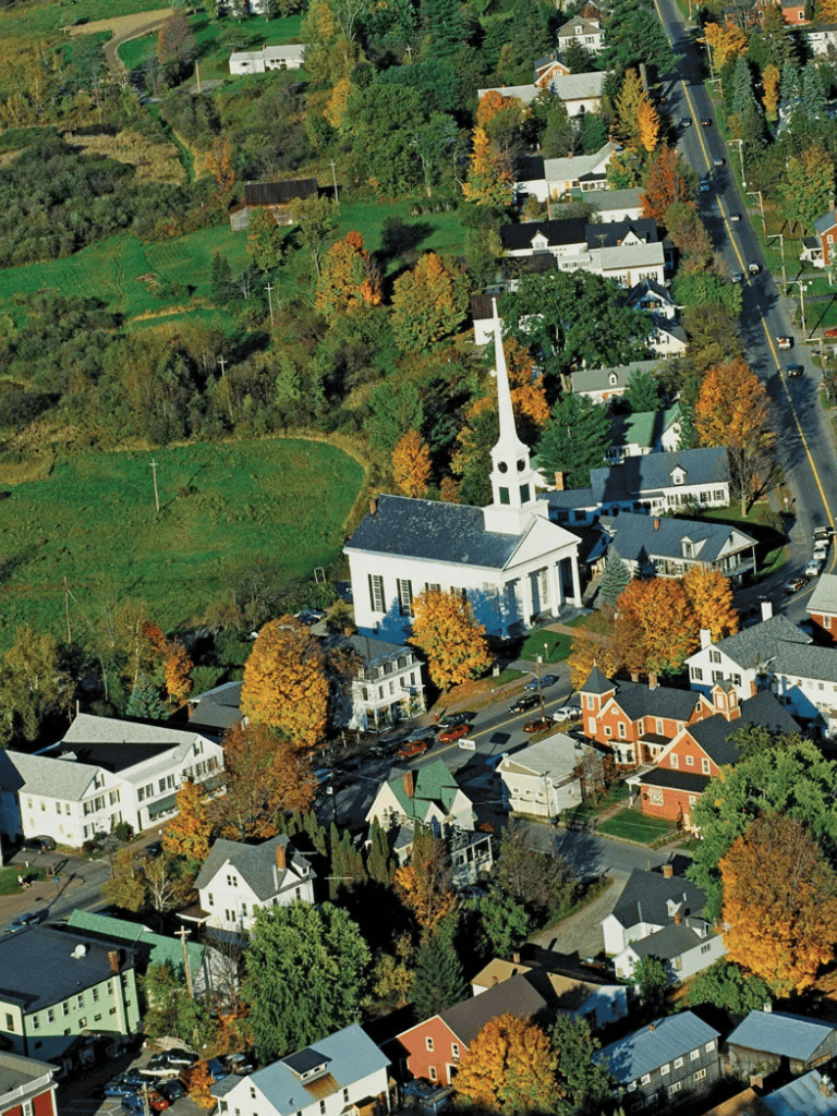 Aerial view of a small town with a prominent white church, colorful autumn trees, and a mix of residential homes and greenery.
