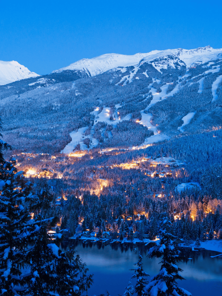 Snowy mountain ski resort at dusk with illuminated slopes and cozy lakeside village.