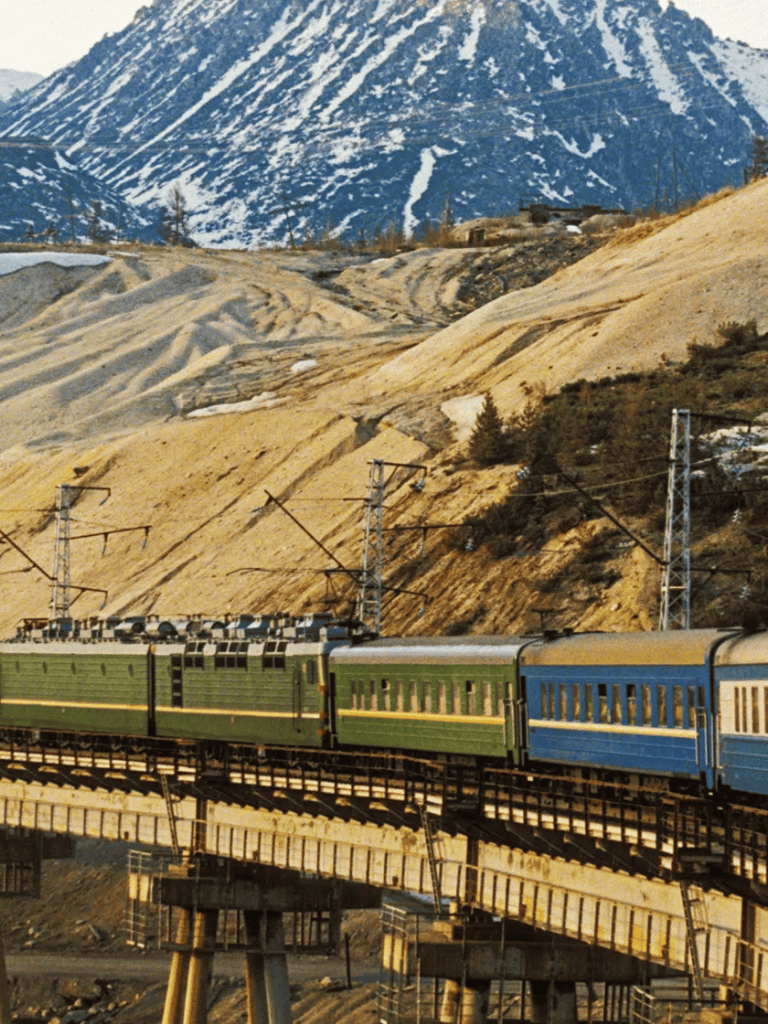 Vibrant green and blue train traveling through mountainous terrain with snow-capped peaks, scenic views, and railway infrastructure.
