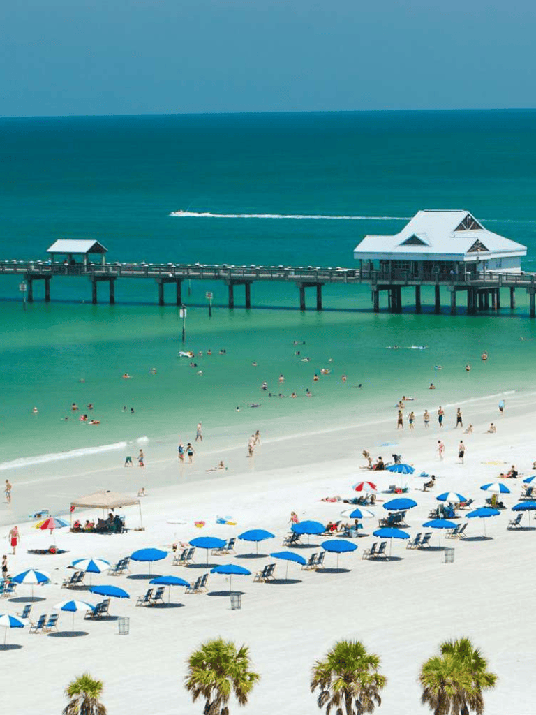Sunny beach with turquoise waters, pier, umbrellas, and beachgoers enjoying the seaside.