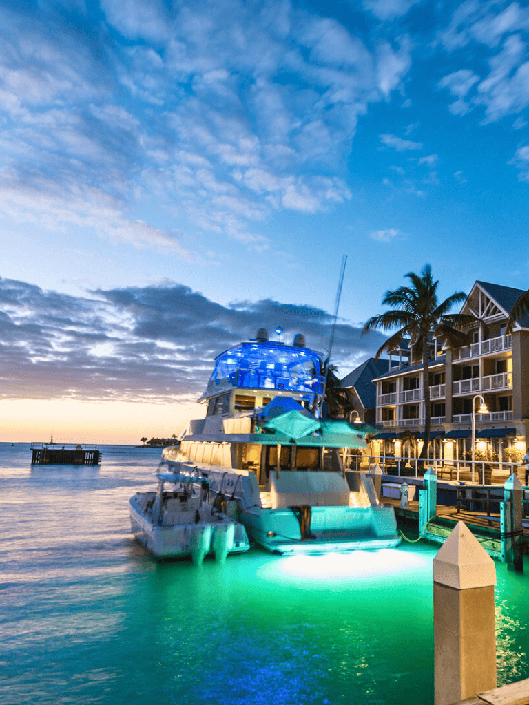 Luxurious yacht docked at tropical marina during sunset with resort buildings and palm trees.