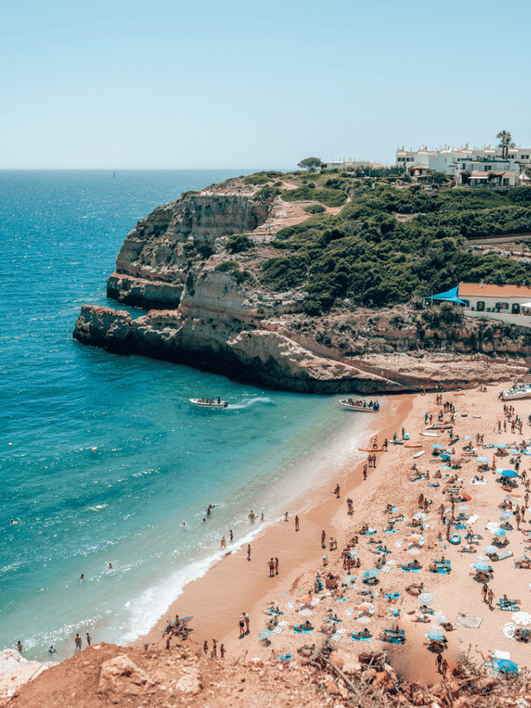 Sunny beach with cliffs and white residential buildings, crowded with umbrellas and visitors.