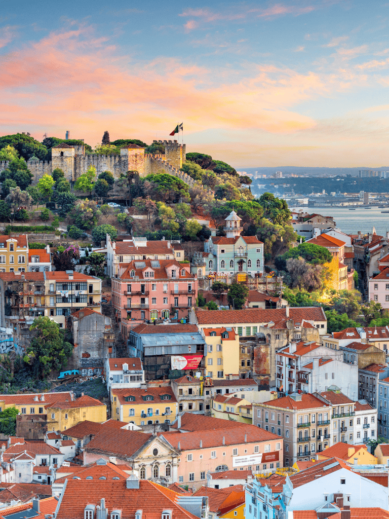 Ancient Lisbon castle overlooking colorful historic neighborhood at sunset near Tagus River.