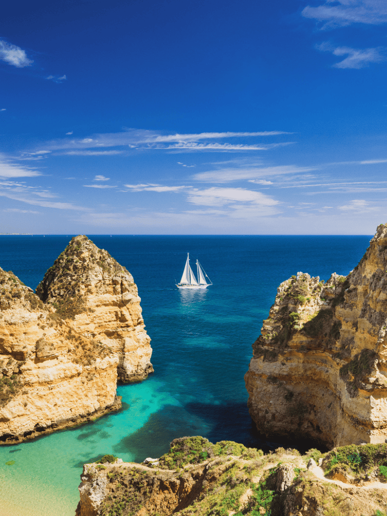 Spectacular coastal cliffs with turquoise waters and a sailboat under blue skies, Portugal.