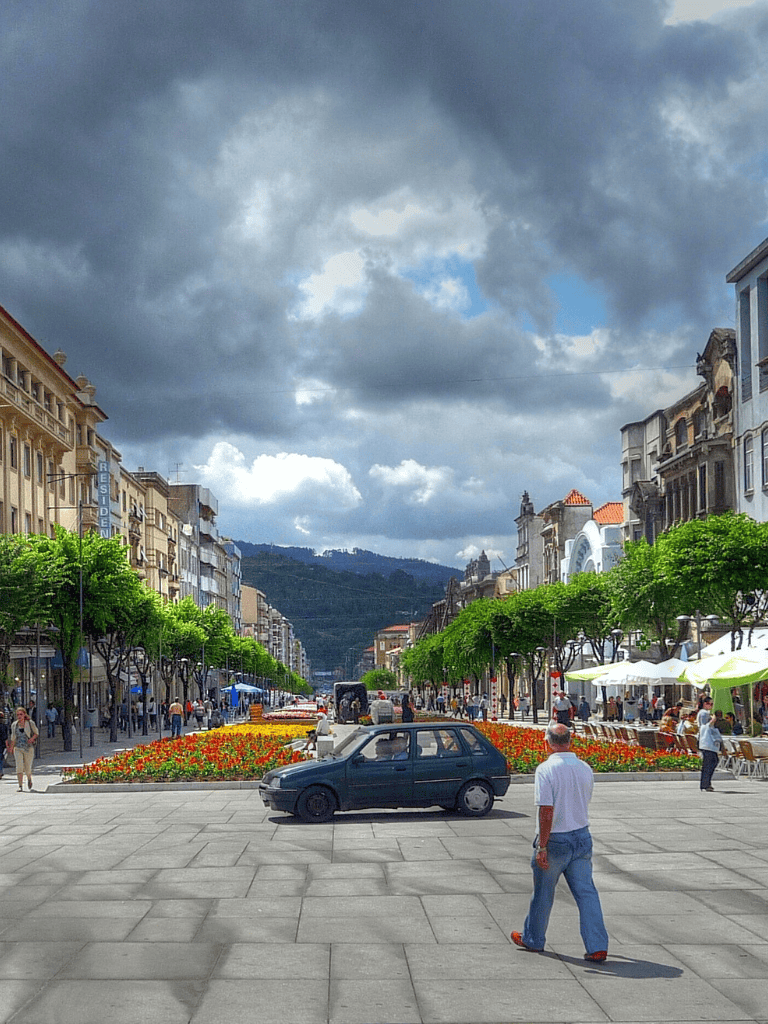 Street view of a bustling city with vibrant flowers, green trees, and historic buildings under cloudy skies.