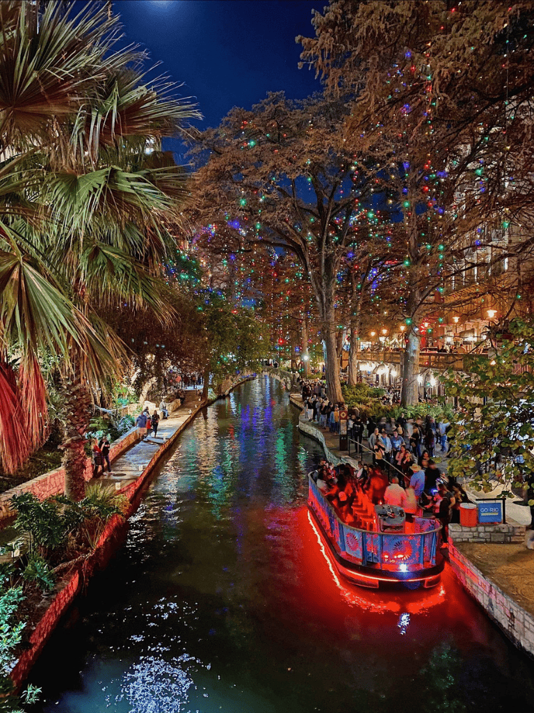 Colorful river walk with boat cruise, lit trees, and lively nightlife in downtown San Antonio.