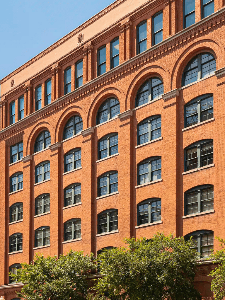 Historic red brick building with large arched windows and lush greenery in front.