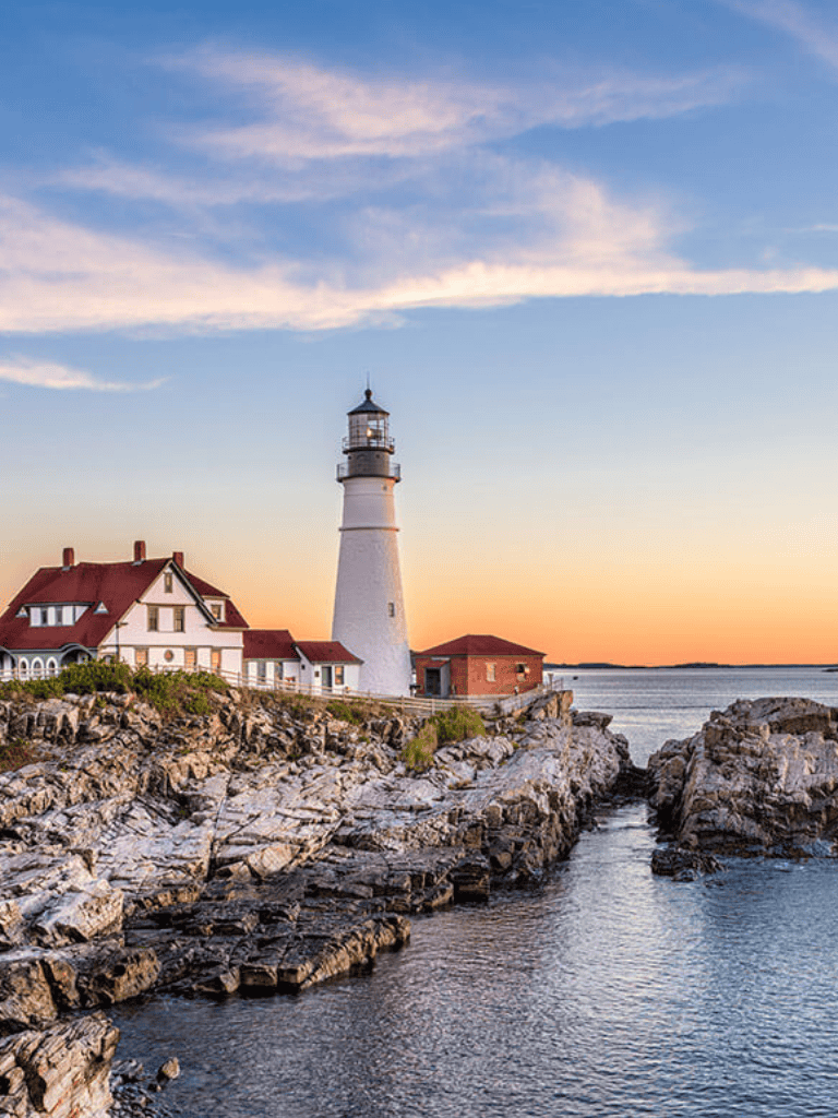 Lighthouse on rocky coast at sunset with blue sky and water, scenic coastal view.