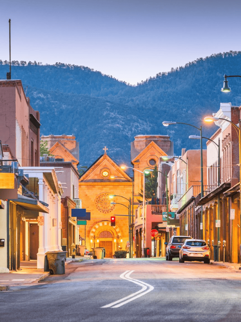 Historic downtown street at dusk with mountain backdrop, featuring shops, streetlights, and parked cars.