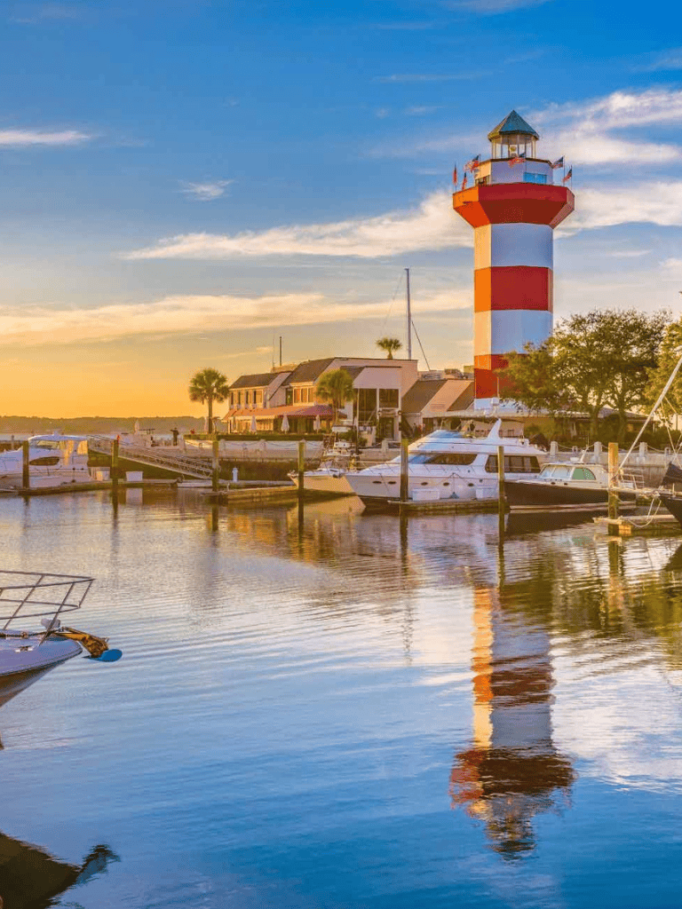 Colorful lighthouse at sunset over marina with boats and waterfront homes, scenic coastal view.