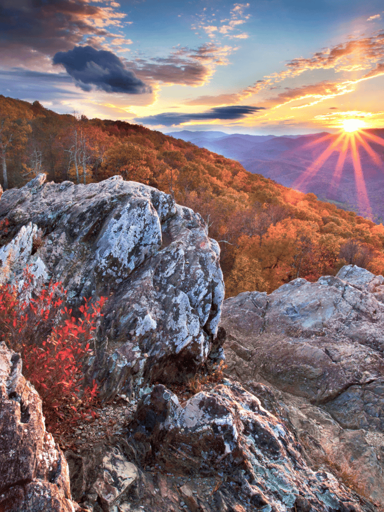 Vast mountain landscape with autumn foliage and dramatic sunset over rugged rocks and colorful trees.