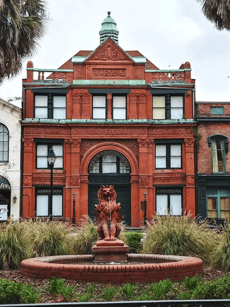 Elegant red brick building with historic architecture, fountain, lion statue, and lush greenery in Savannah.