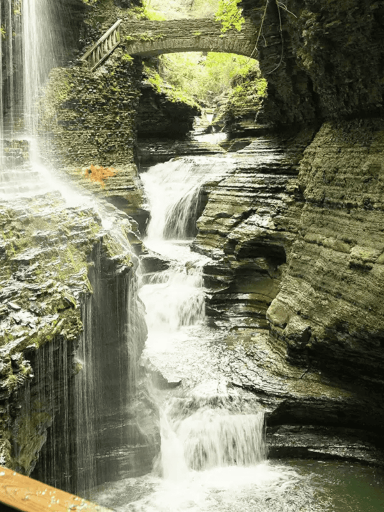 Serene waterfall in a lush forest with a stone bridge over flowing water.