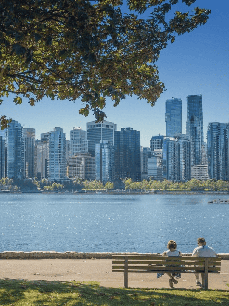 Aerial view of modern city skyline across a river with people enjoying a park bench.