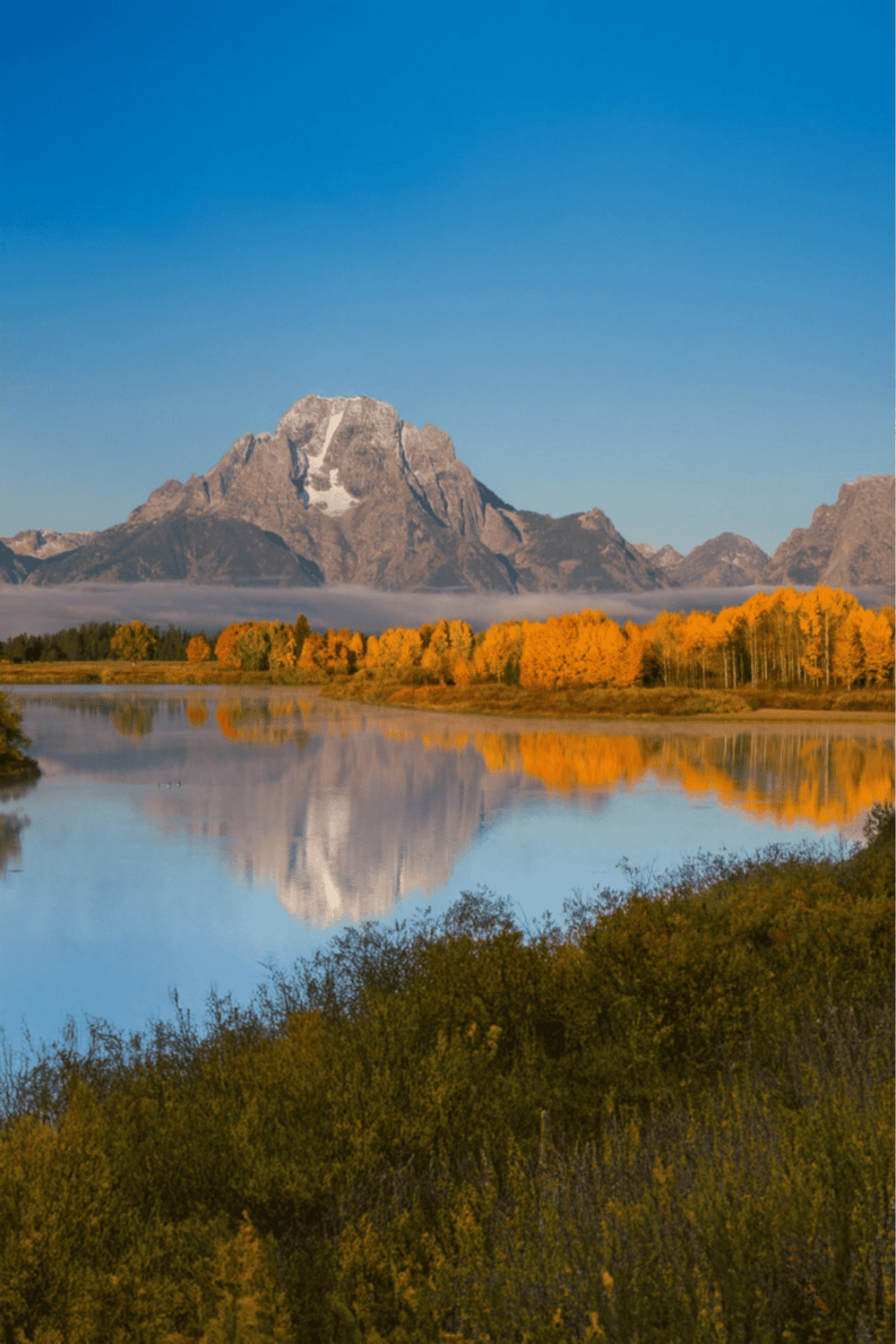 Majestic mountain landscape with reflection in calm water, vibrant fall foliage, and clear blue sky.